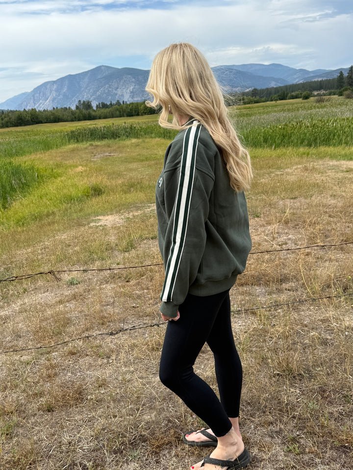 Person standing wearing a green Montana crewneck sweatshirt with a scenic background of mountains and fields.