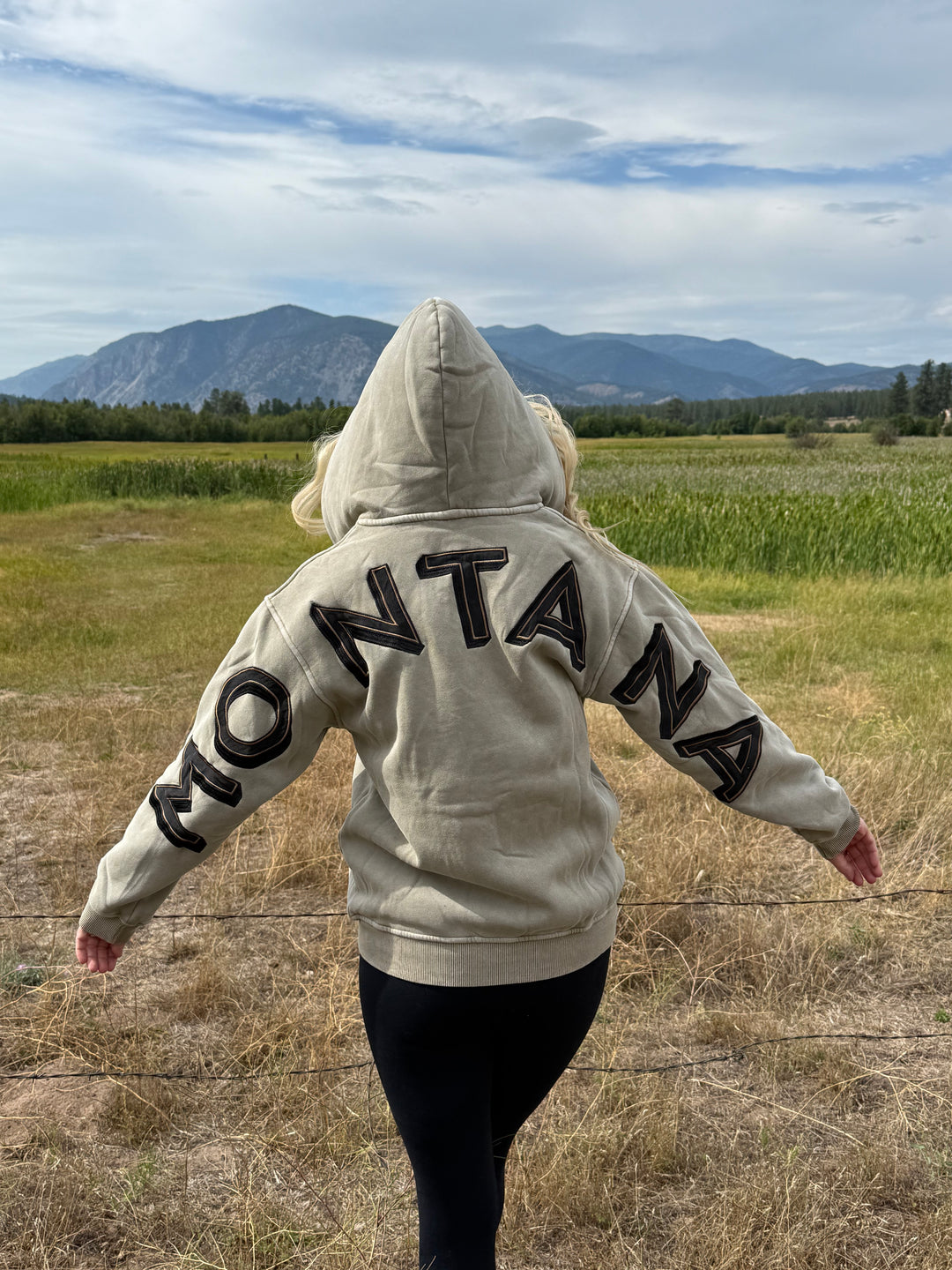 Person wearing a hoodie with MONTANA text in an open field with mountains in the background