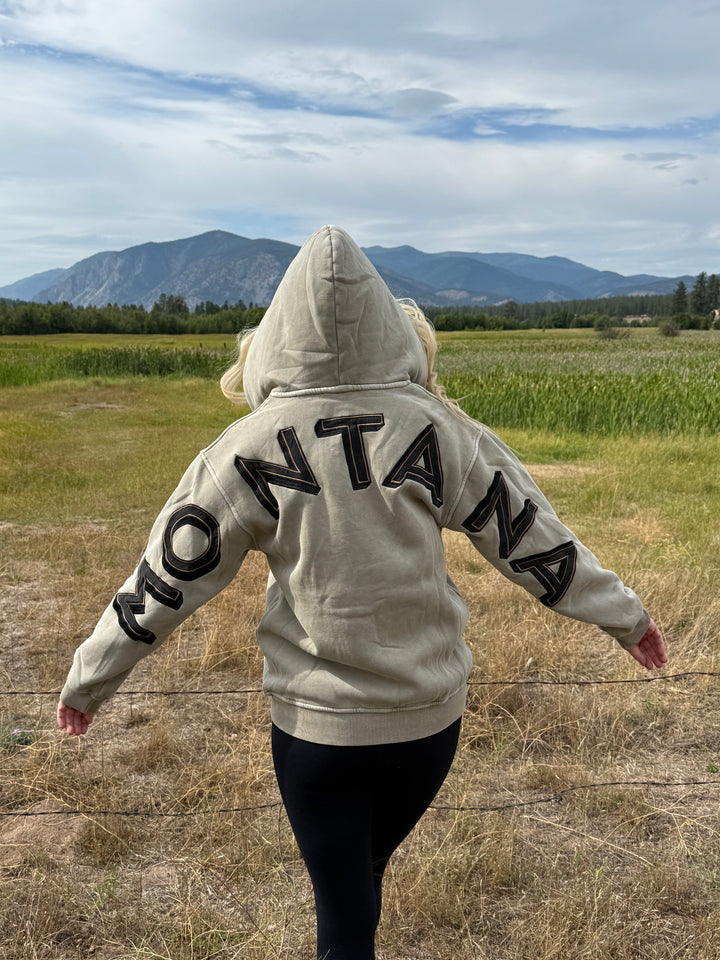 Person wearing a hoodie with MONTANA text in an open field with mountains in the background