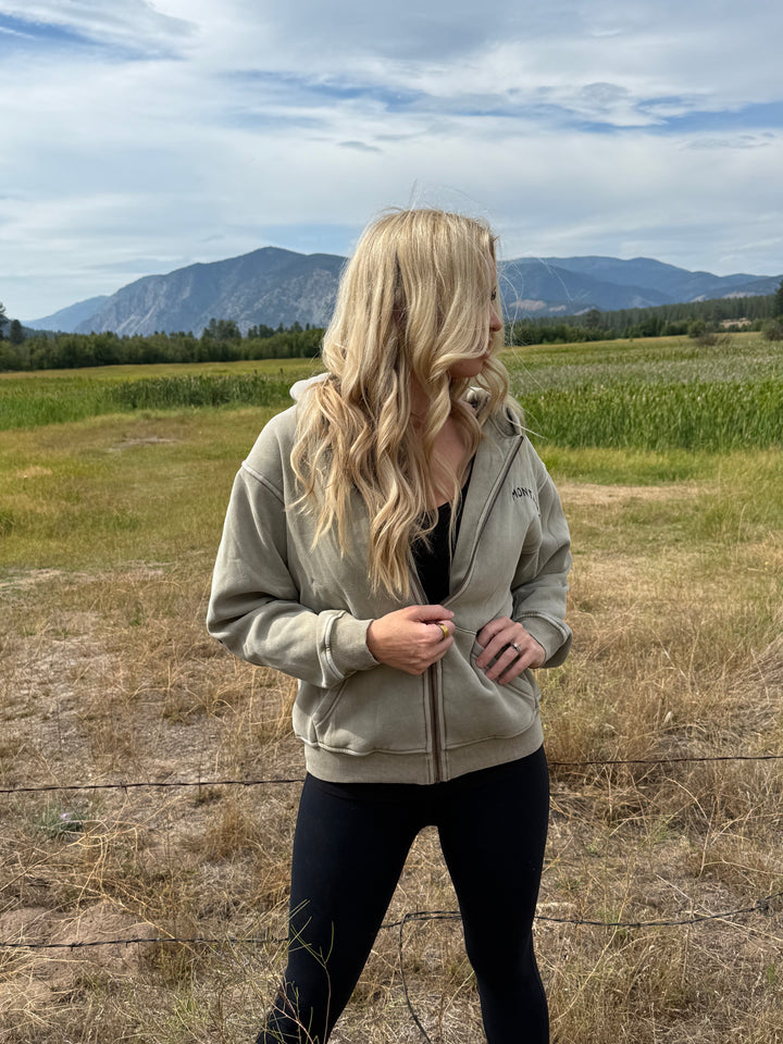 Person wearing an olive Montana Zip hoodie standing in a field with mountains in the background
