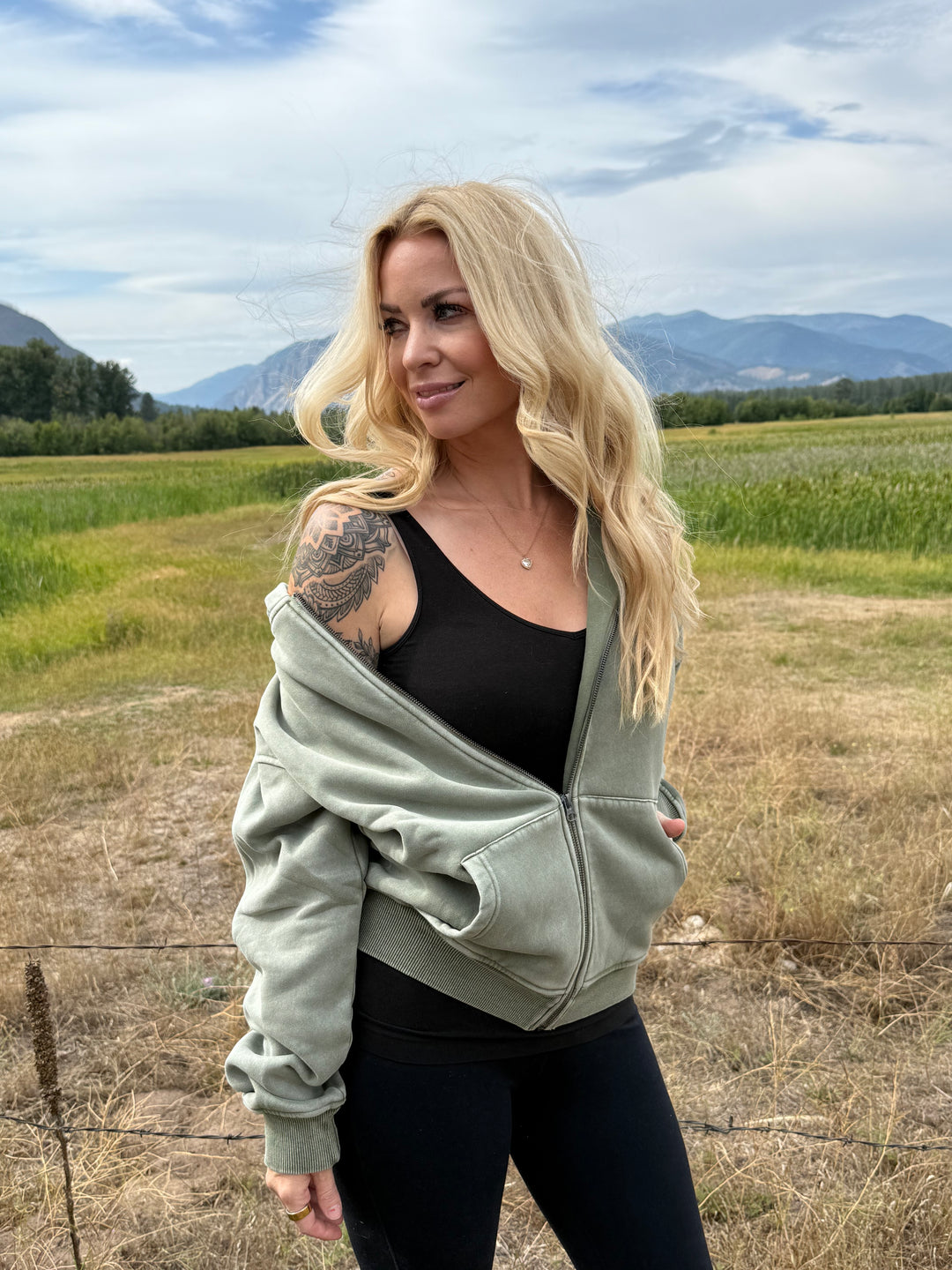 Person wearing a green Montana Zip hoodie over a black top in a field with mountains in the background