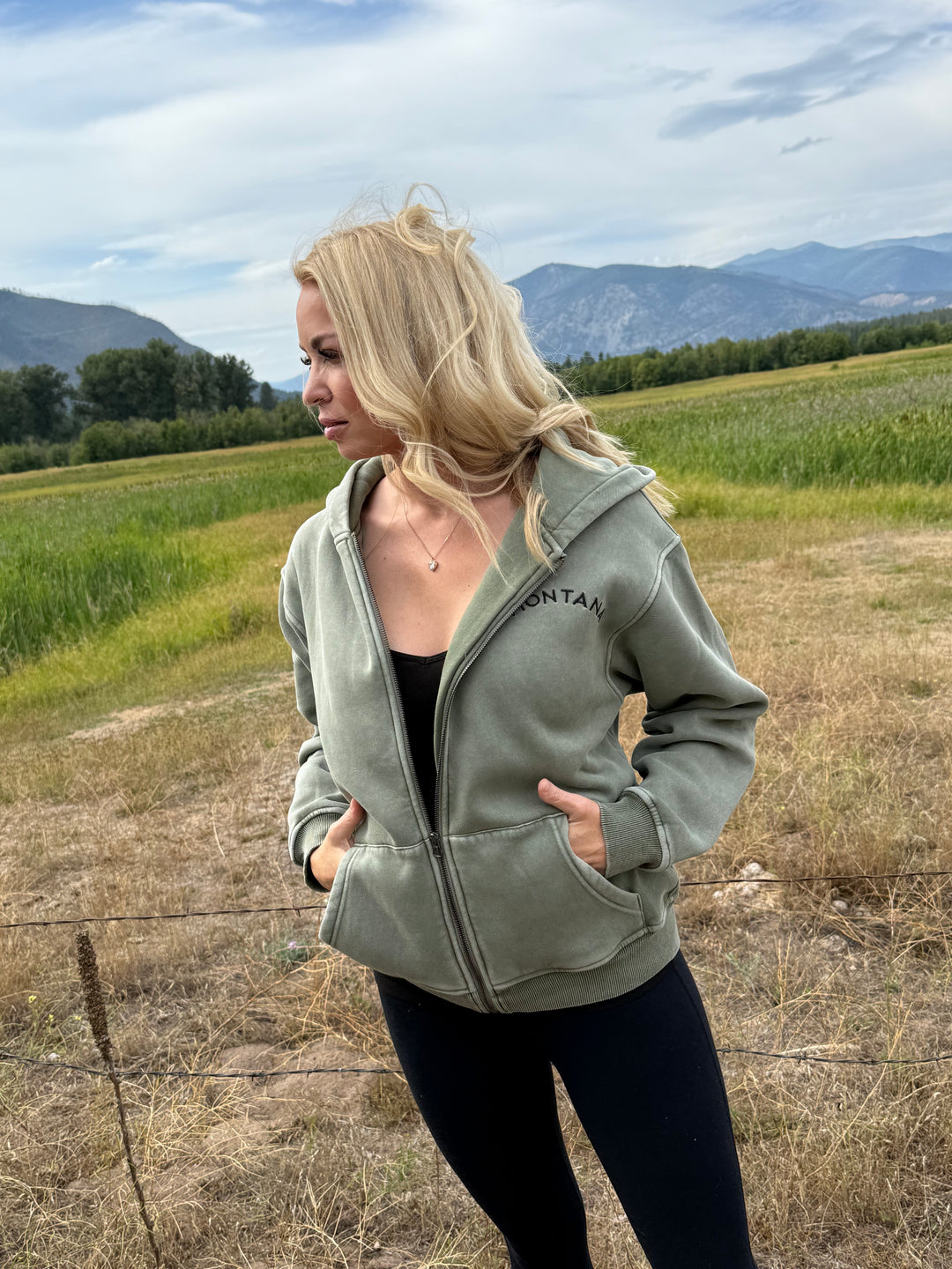 Person wearing a green Montana zip hoodie standing in a field with a mountainous background