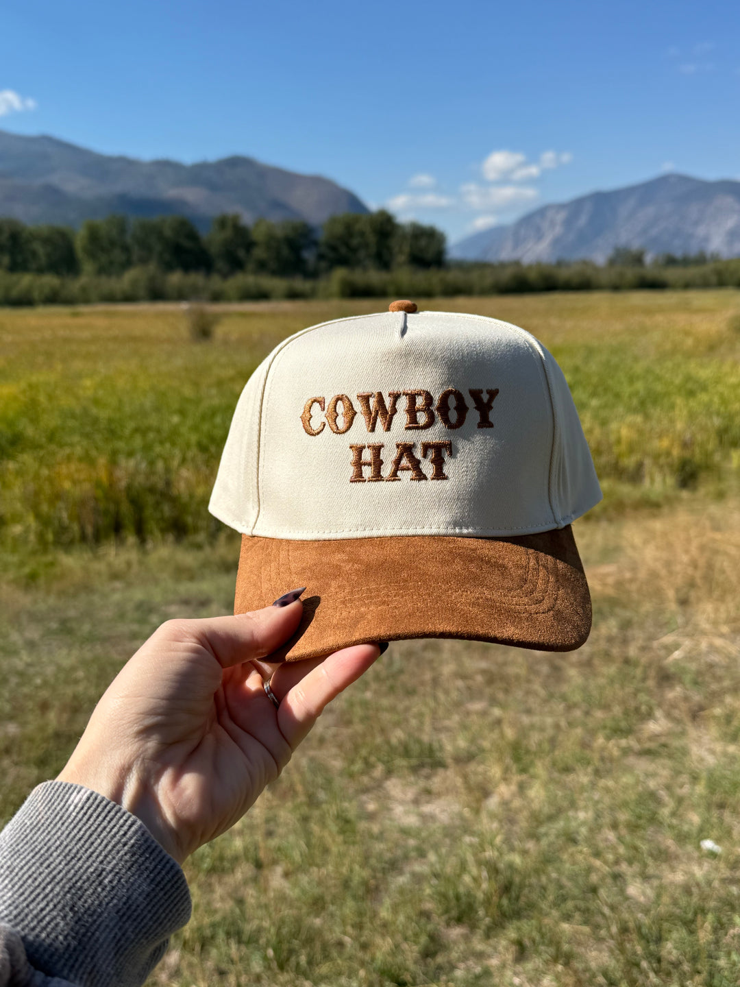 Hand holding a trucker hat with 'Cowboy Hat' text against a scenic background
