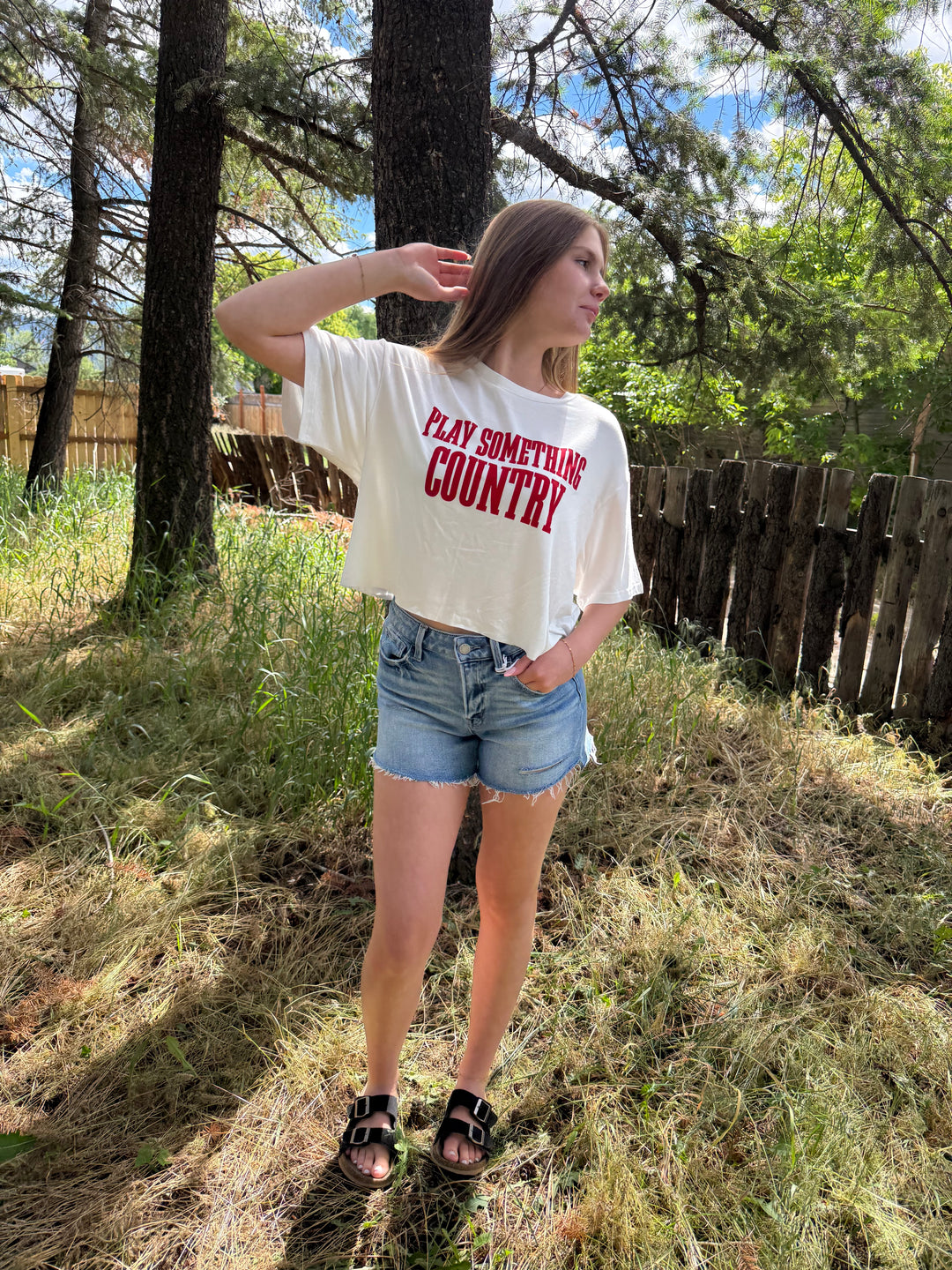 Person wearing a 'Play Something Country' t-shirt in a natural setting with trees and grass.