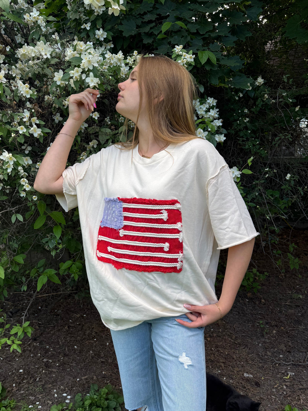 Person wearing a white t-shirt with an American flag design, standing outdoors near greenery.